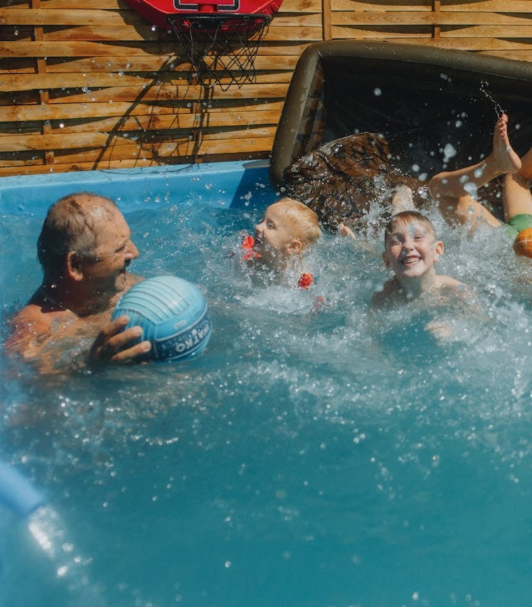 Grandfather and grandchildren playing in a small pool with a basketball hoop in the background.