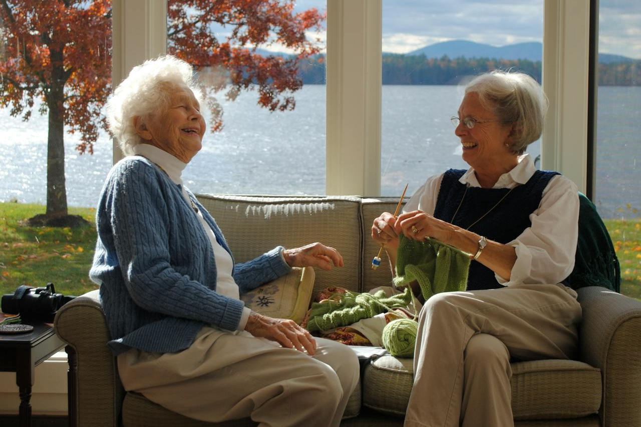 Two elderly women sitting on a couch by a large window with a scenic view of water and trees.