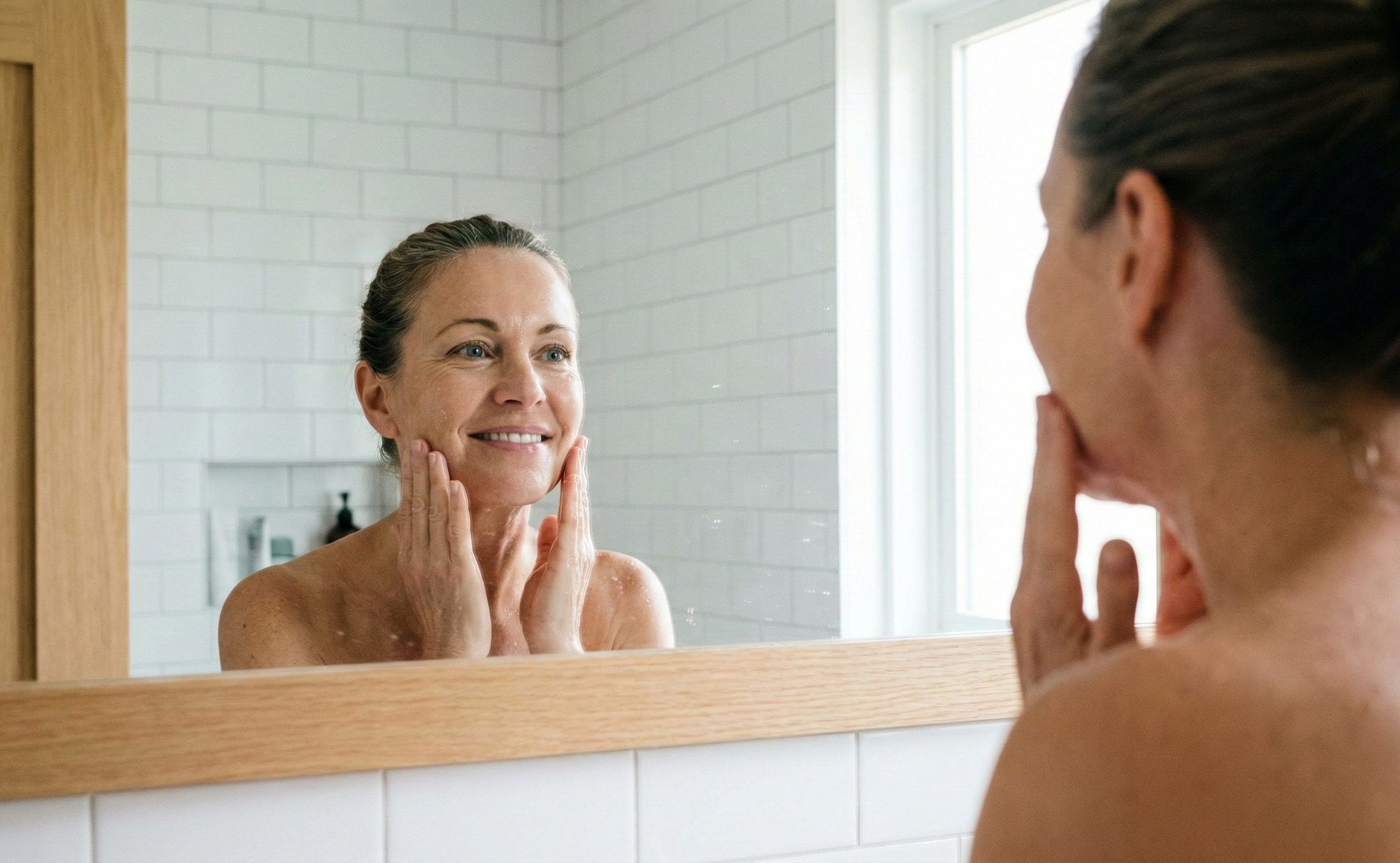 Woman with clear, healthy skin, looking radiant in front of a bathroom mirror.